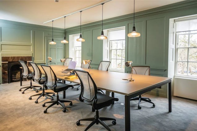 Mesh chairs surround a wooden table in a room with green panelled walls, pendant lights, and a fireplace.