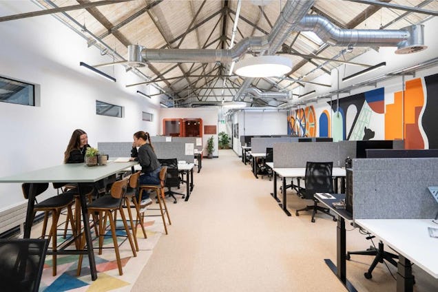 People sat at high tables while others work at desks in an office with metal ceiling pipes.