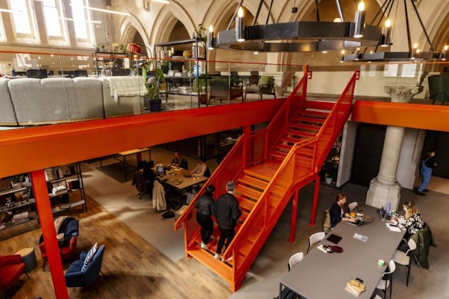 People walk on an orange staircase and sit at desks in a coworking space with arches and pillars.