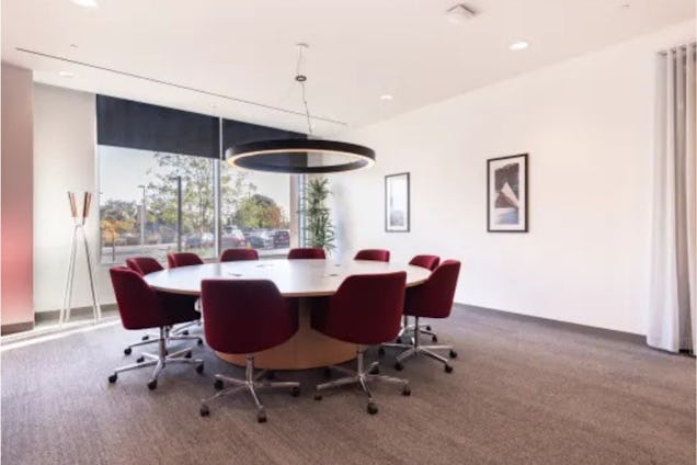 Red chairs surround a circular wooden table in a meeting room with a pendant light and carpet.