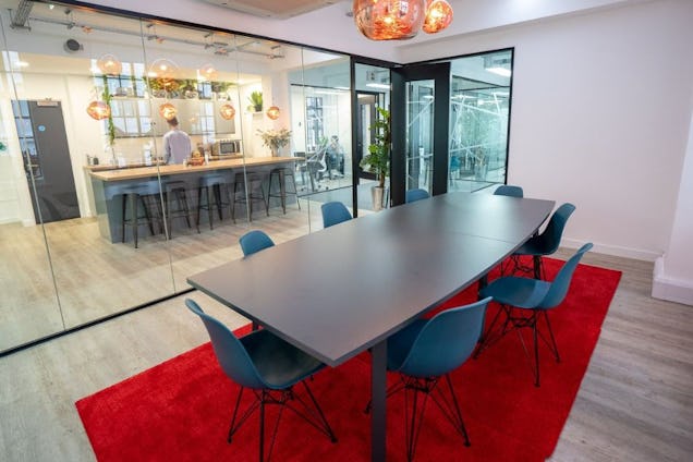 Blue chairs surround a table on a red rug in a room with a person standing at a kitchen bar.