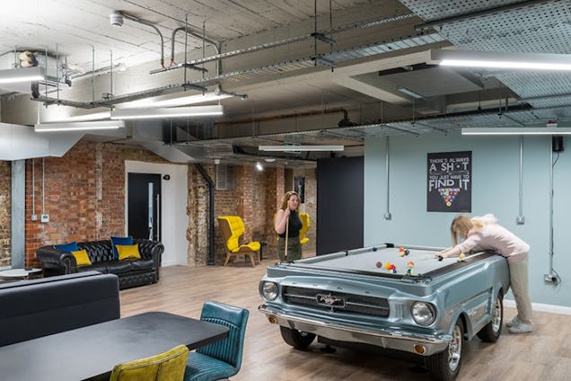 A person plays pool on a table shaped like a car in a lounge area with sofas and brick walls.