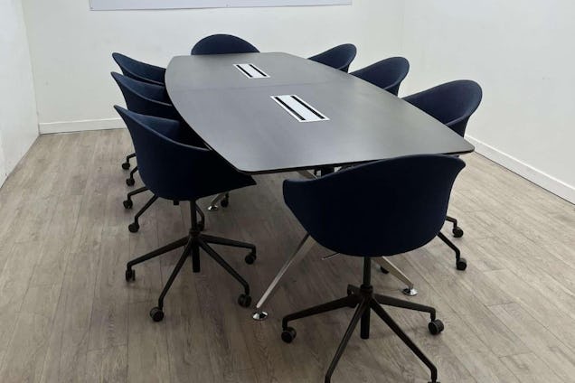 Black chairs with wheels surround a rounded table on a wooden floor in a room with white walls.