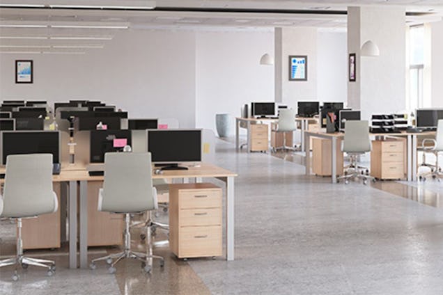 Rows of wooden desks with white chairs and computer monitors fill an open-plan office.