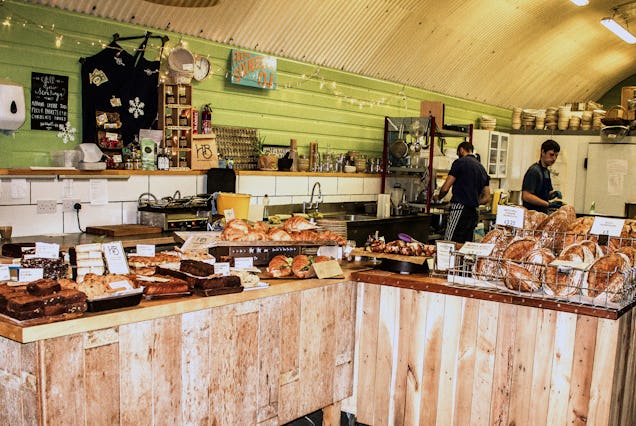 Trays of pastries sit on wooden counters in a bakery while staff members work behind the counter.