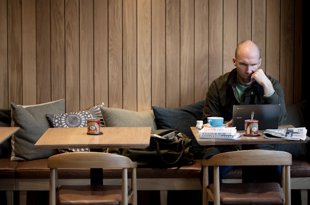 A person at a wooden table using a laptop on a bench with cushions in front of a wood-panelled wall.