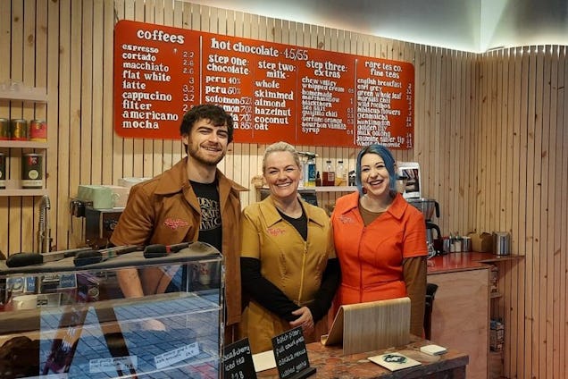 Three staff members stand behind a counter and smile in front of a menu board and wood-panelled wall.