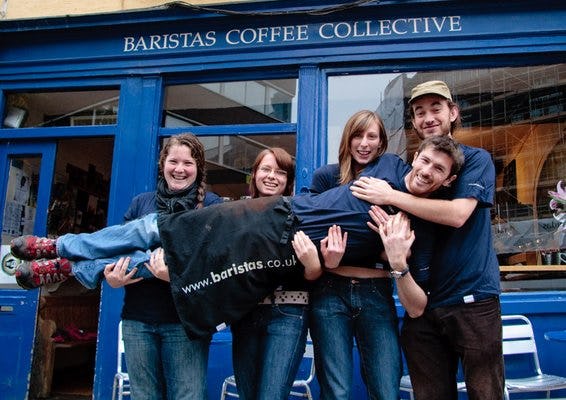 Four people pose and smile for a photo in front of a blue storefront with a "BARISTAS COFFEE COLLECTIVE" sign.