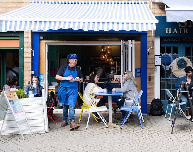 A person in a blue apron stands near customers sitting at a table under a striped awning.