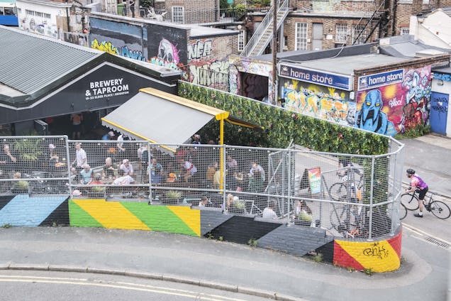 People sat at tables in an outdoor seating area with a fence and awning next to buildings with graffiti and a person on a bicycle.