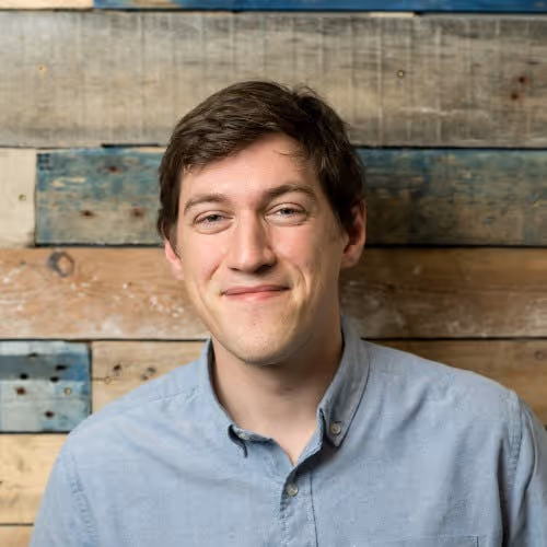 Man in a light grey shirt stood against a rustic wood effect wall.
