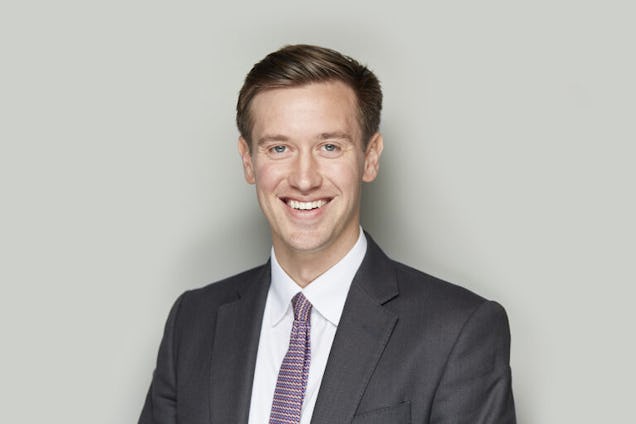 A professional headshot of a smiling man in a dark grey suit and patterned tie against a grey background.