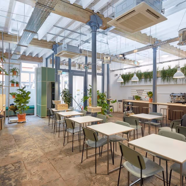 Bright communal workspace with long tables, hanging plants and industrial-style ceiling under a glass roof.