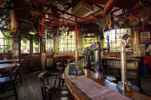 Traditional pub interior with wooden bar, stools and eclectic décor, featuring hanging signs and a cosy atmosphere.