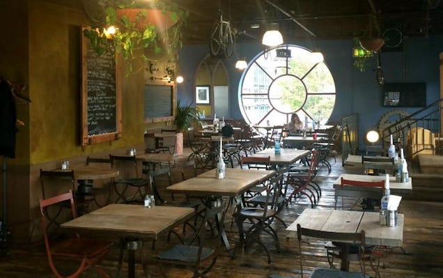 Cafe interior with square wooden tables, bistro chairs, hanging vines, and a distinctive large circular window.