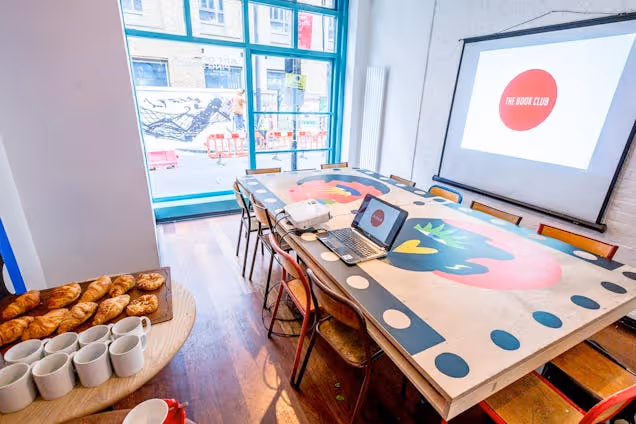 Bright meeting room with a colourful table, screen for presentations and pastries with coffee set out for attendees.