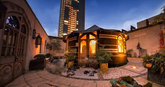 Outdoor courtyard with a lit wooden cabin, seating and plants at dusk, set against surrounding buildings.