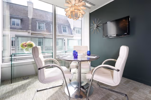 Small meeting area with two white chairs and a round table by large windows, featuring modern décor and city views.