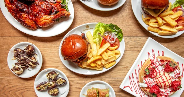Top-down view of a wooden table spread featuring burgers with fries, roasted chicken, small dessert plates, and a strawberry waffle.
