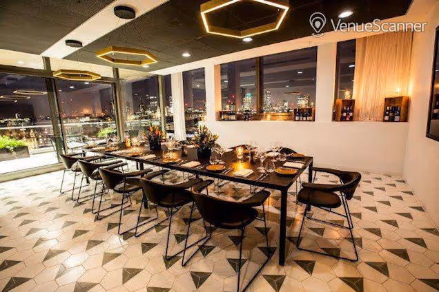 Restaurant interior with a long dining table, patterned floor, warm lighting and large windows overlooking the city.