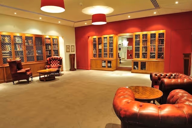 Warm lounge with red walls, leather seating and shelves of wine bottles, featuring tables and soft lighting.