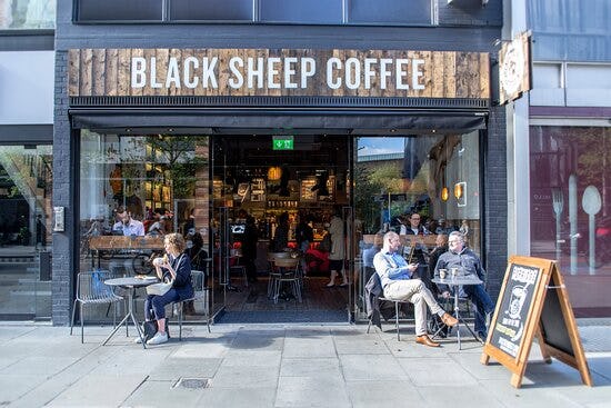 Customers sitting outside Black Sheep Coffee, with people inside the café and a pavement sign at the entrance.