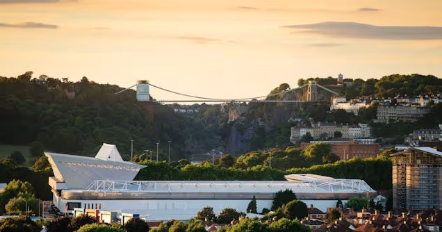 View of Clifton Suspension Bridge over a green valley with buildings and a waterfront area in the foreground at sunset.