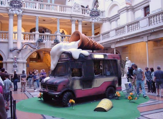 Large ice-cream sculpture draped over a small vehicle in a busy courtyard, with people gathered around the display.