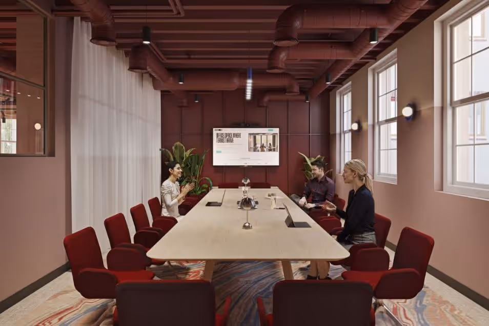 Boardroom with a long light table, red chairs, maroon walls, and an industrial maroon ceiling with exposed pipes.