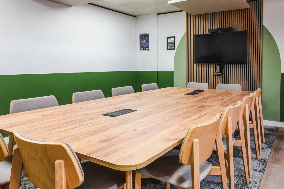 Meeting room with a large light-wood table, wooden chairs, a green accent wall, and a TV mounted on a slatted wood panel.
