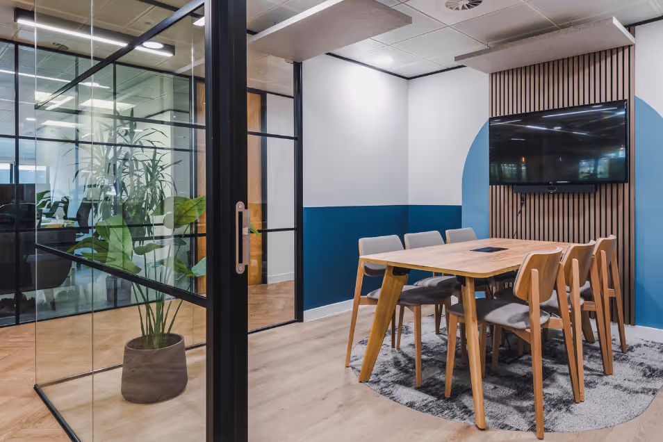 Meeting room with wood furniture and blue accent walls, viewed through a black-framed glass partition with a large potted plant.