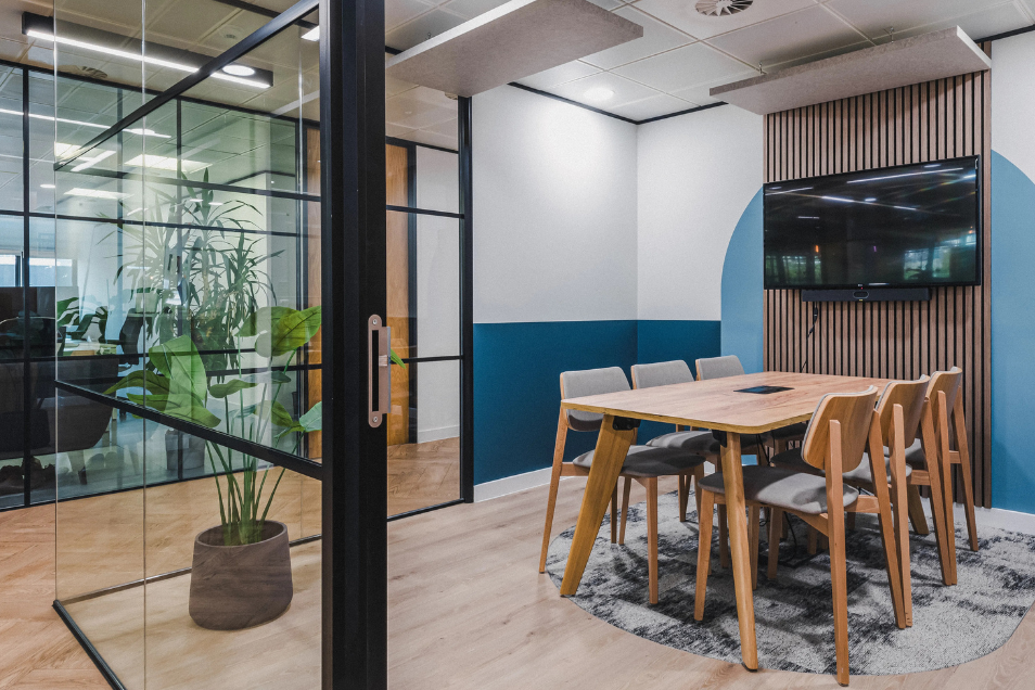 Meeting room with wood furniture and blue accent walls, viewed through a black-framed glass partition with a large potted plant.