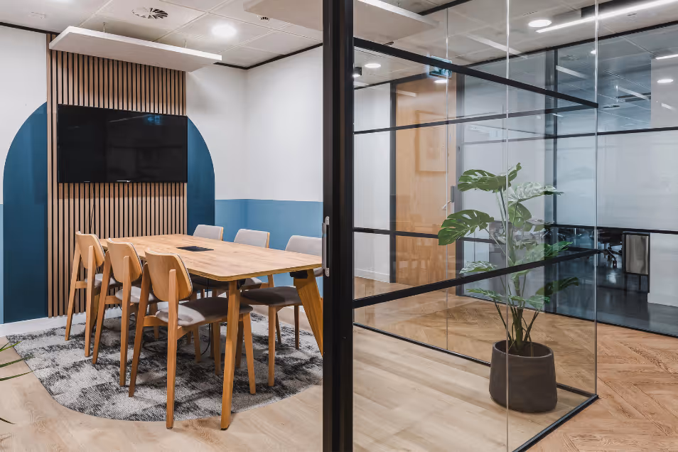 Meeting room with wood furniture and blue walls, viewed through a black-framed glass partition beside a large indoor plant.