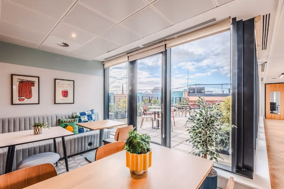 Sunlit office break room with wooden tables and a cushioned bench, featuring large glass doors that open onto an outdoor terrace.