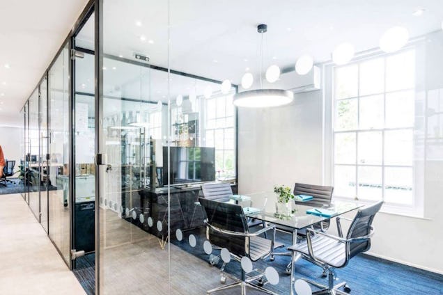 Meeting room with a glass table, black mesh chairs, and large white-framed windows, separated by glass partitions.