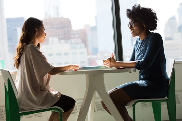 Two women sit at a small white table for a meeting in a bright room with a city skyline visible through the window.