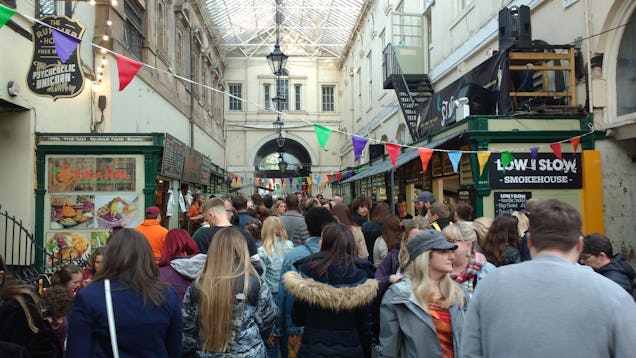 Busy indoor market with crowds of people, colourful bunting and food stalls under a covered arcade.