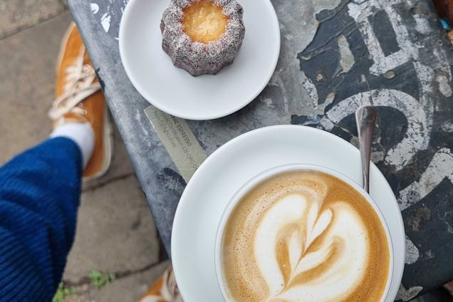 Cup of coffee with latte art beside a pastry on a café table, with a person’s legs and shoes visible below.