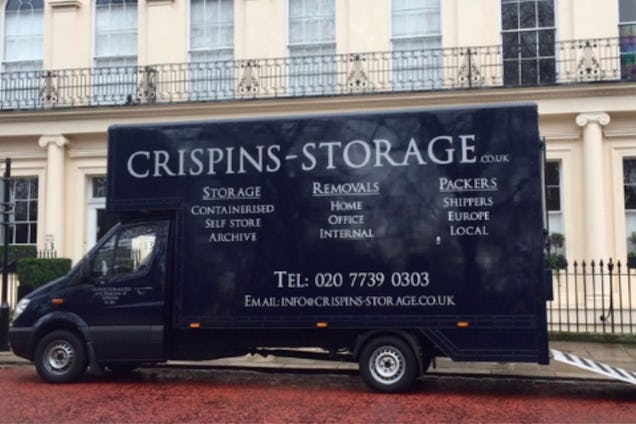 A large black Crispins Storage removal truck with white lettering and a rear ramp is parked on a city street.