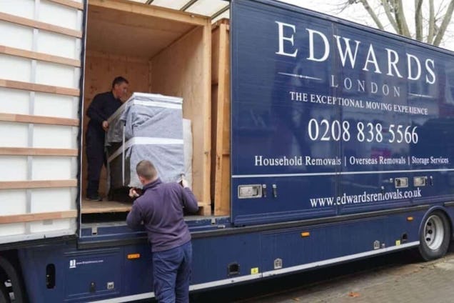 Two movers carefully maneuver a large, wrapped piece of furniture into the back of a blue Edwards London removal truck.