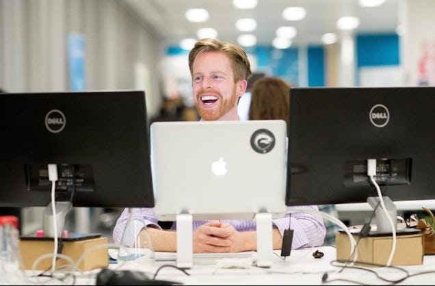 A smiling man with a beard sits at his desk with a laptop, while a colleague works in the background of a bright, modern office.