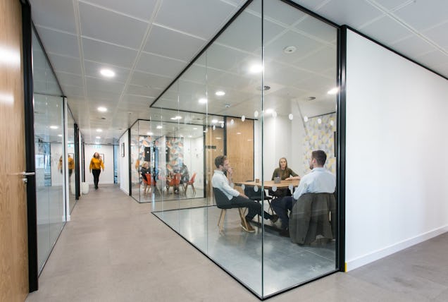 Office corridor lined with glass-walled meeting rooms, where teams are visible collaborating at wooden tables under bright lighting.