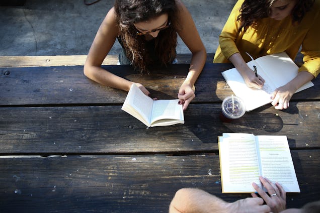 High-angle view of three people sitting at a rustic wooden table, focused on writing and reading in their notebooks.