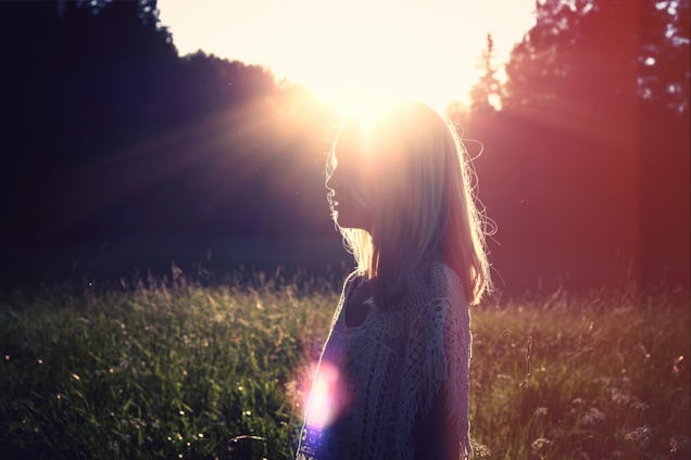 A woman stands in a grassy field, silhouetted against the glowing rays of a setting sun.