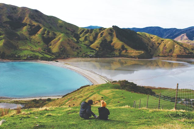 Two people sat on a grassy hilltop, looking out over a blue bay and green coastal mountains.