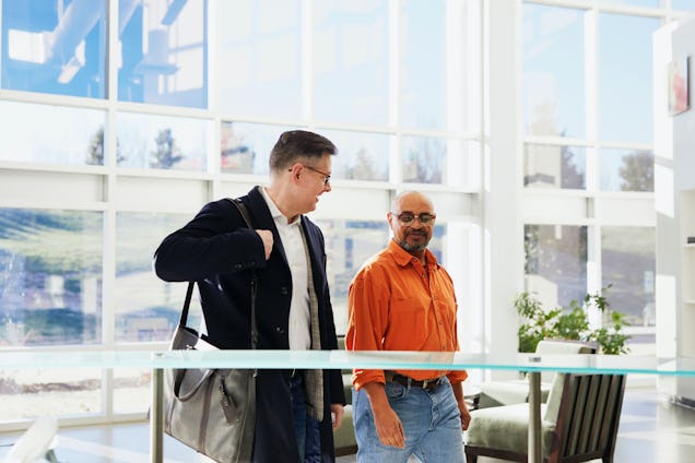 Two people walk and talking at the entrance with glass walls and plants.