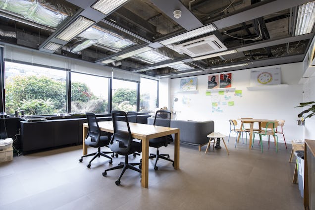 Tables and chairs sit in an office workspace with windows, a sofa, and exposed ceiling pipes.