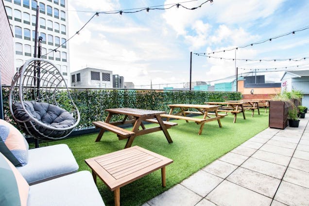 A sunny rooftop terrace with artificial grass, wooden picnic tables, a hanging egg chair, and string lights.