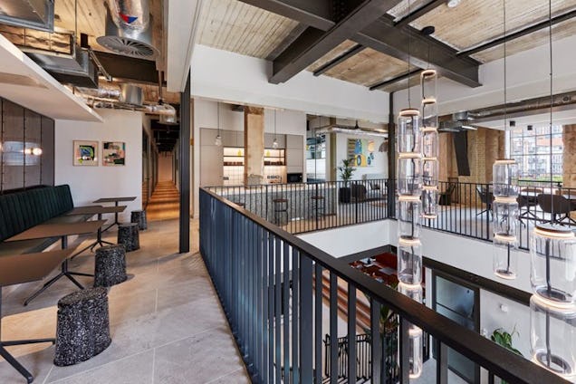 A multi-level industrial office balcony with black railings overlooking a bright, open workspace with hanging lights.