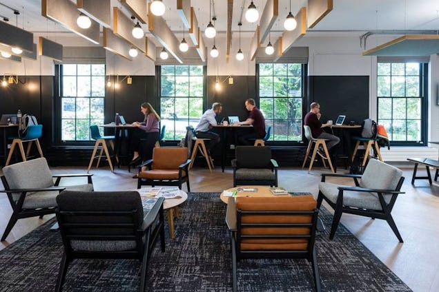 Professionals work at communal tables in a bright, modern lounge with grey armchairs and a decorative wood-slat ceiling.
